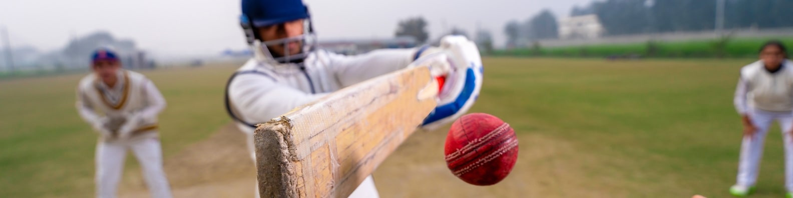 A cricket batsman in protective gear swings his bat to hit a red ball on a grassy field, with two fielders blurred in the background. Streaming Cricket Blog illustration